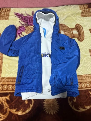 Blå pufferjacka från Nike med huva - Säljer en blå pufferjacka från Nike med huva och svarta detaljer. Jackan har dragkedja framtill, två sidofickor med dragkedja och ett Nike-märke på bröstet. Materialet är glansigt och syntetiskt, perfekt för kyliga dagar. Jackan är långärmad och har elastiska muddar vid ärmslut och nederkant.