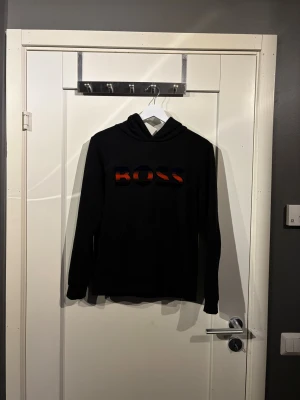 Svart hoodie från BOSS - Svart hoodie från BOSS med stor, broderad logga i blått och orange på bröstet. Tröjan har huva, magficka och långa ärmar. Perfekt för dig som gillar stilren streetwear med en touch av färg. Litet märke bak på ryggen.