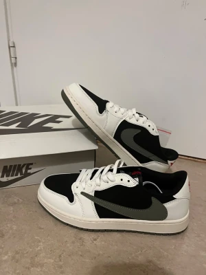 Nike Jordan 1 Low svart/vit/grå - Nike Jordan 1 Low sneakers med svart ovandel, vita paneler och grå swoosh. Klassisk rund tå, snörning och platt sula i grått. Ikonisk design med kontrasterande färger och stilren look. Perfekt för dig som gillar streetwear och vill sticka ut med snygga sneakers.