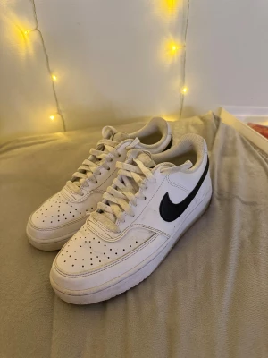 NIKE SNEAKER COURT VISION LÅG NEXT NATURE - VIT/SVART - Fräscha Nike sneaker court vision låg next nature i vitt med svart swoosh på sidan. Klassisk låg modell med snörning, perforerad tå och platt sula. Skorna är tillverkade i skinn och har rund tå. Perfekt för dig som gillar streetstyle och vill ha en ikonisk sneaker i garderoben. Köptes för 1000kr vid riktigt köp intresse kan jag tänka mig att sänka priset.