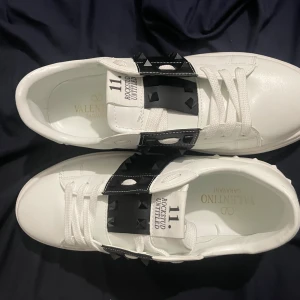 Valentino Rockstud Untitled sneakers - Snygga vita Valentino Rockstud Untitled sneakers med svarta detaljer och ikoniska pyramidnitar. Skorna har rund tå, platt sula och snörning. Tillverkade i skinn med en cool kontrast mellan vitt och svart. Perfekt för dig som gillar lyxiga och edgy sneakers.