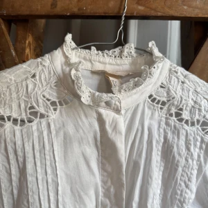 Vit blus med spetsdetaljer  - Vit blus / skjorta med fina spetsdetaljer vid axlar och ärmar, samt dekorativ krage med broderad spets. Blusen har knappar framtill och är långärmad med lätt puff vid axlarna. 