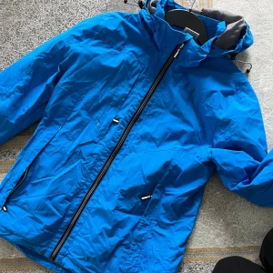 Blå vindjacka från Riva, storlek M - Blå vindjacka med huva från Riva, perfekt för blåsiga dagar. Jackan har svarta detaljer, dragkedja framtill och två sidofickor med dragkedja. Insidan är grå och materialet känns lätt och smidigt. Justerbar huva med snörning för extra skydd.