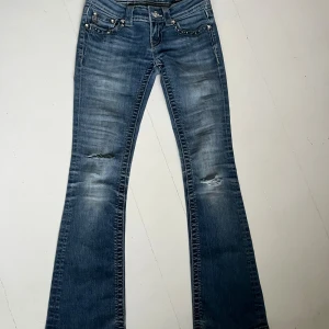 Miss Me blå bootcut jeans med nitar - Snygga blå jeans från Miss Me med bootcut passform. Jeansen har slitningar vid knäna och coola detaljer med nitar och strass på fickorna. Låg midja och klassisk femficksmodell. Perfekta för dig som gillar statement-jeans med extra bling.