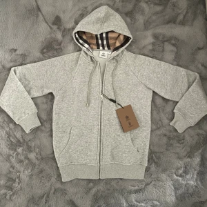 Grå hoodie från Burberry  - Storlek:XS.                                                    Passform/kvalite:Helt ny,passar perfekt för folk som vill att den ska sitta lite tight.Funkar även om du bär en storlek mindre alltså 158-164.                                Beskrivning:Snygg grå hoodie från Burberry med klassiskt rutigt foder i huvan och broderad logga på bröstet. Tröjan har dragkedja framtill, snörning i huvan och två fickor. Perfekt för dig som vill ha en stilren och lyxig look.