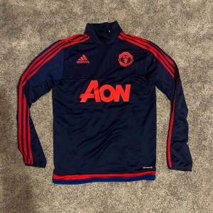 Manchester United långärmad träningströja från Adidas - Mörkblå Manchester United träningströja från Adidas med röda detaljer och loggor. Långärmad modell i funktionsmaterial (climacool) med hög krage, röda ränder längs ärmarna och stor AON-logga på bröstet. Perfekt för dig som älskar fotboll och vill visa att man är en riktig fan!