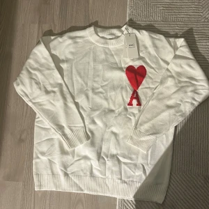 Vit stickad tröja från Ami - Vit stickad tröja från Ami Alexandre Mattiussi med rund hals och ribbade muddar. På bröstet finns ett stort rött hjärta och bokstaven A som broderad detalj. Perfekt för dig som gillar stilrena plagg med en twist.