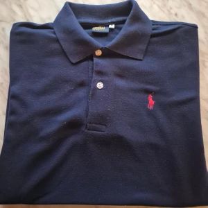 pikétröja  Ralph Lauren - Klassisk marinblå pikétröja från Ralph Lauren med korta ärmar och två vita knappar framtill. Skick : 9/10