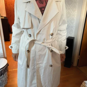 Beige trenchcoat med bälte - Snygg beige trenchcoat med klassisk dubbelknäppning och bälte i midjan. Jackan har stora kragar, långa ärmar och dekorativa knappar. Perfekt för dig som gillar stilrena och tidlösa plagg.