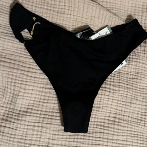 Svart bikiniunderdel med gulddetalj - Svart bikiniunderdel från H&M med hög midja och en snygg guldfärgad metall-detalj på ena sidan. 
