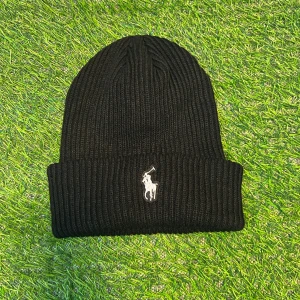 Svart ribbad mössa från Polo Ralph Lauren - Svart ribbstickad mössa från Polo Ralph Lauren med klassisk broderad logga i vitt framtill. Mjuk och varm, perfekt för kyliga dagar. Stilren design med uppvikt kant som ger en snygg look.