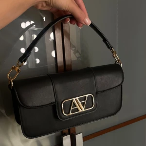 Svart axelväska - Snygg svart axelväska i skinn med guldfärgad metallogo framtill. Väskan har en stilren, rektangulär form och ett kort axelband med gulddetaljer. Perfekt för dig som gillar minimalistisk design och exklusiva detaljer.