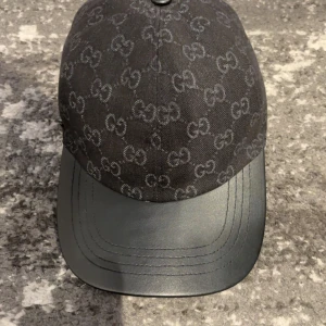 Svart Gucci keps med GG-mönster - Snygg svart keps från Gucci med klassiskt GG-mönster över hela kepsen. Skärmen är i svart skinn och kepsen har justerbar rem baktill. Perfekt accessoar för dig som vill sticka ut med en lyxig och trendig look. Helt oanvänd eftersom kepsen inte passade mitt huvudform 