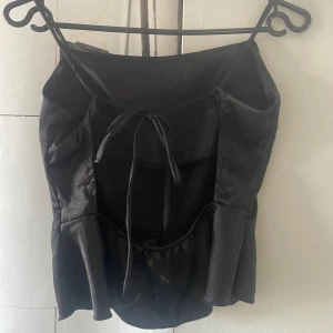 Svart satinlinnetopp med knyt - Snygg svart linne-topp i satin med smala axelband och trendig knytning framtill. Toppen har volangdetaljer nedtill och en stilren, slät baksida. Perfekt för dig som gillar en chic och stilren look.