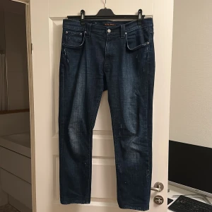 Mörkblå jeans från Nudie Jeans - Lean dean. 34w sitttr som 33w, 34l kortat ned de så de är 30l. Snygga mörkblå jeans från Nudie Jeans med klassisk femficksdesign och subtila slitningar på fram- och baksida. Jeansen har raka ben och silverfärgade nitar vid fickorna. Materialet är robust denim i bomull med en cool tvättad look.  Inga defekter. Priset är inte hugget i sten!