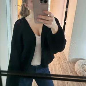 Svart stickad kofta - Säljer en svart stickad kofta med ballongärmar och lös passform. Koftan är öppen framtill och har ett luftigt, grovstickat material som ger en chill vibe. Perfekt att slänga över ett linne eller t-shirt för en avslappnad look. Använd 1 gång