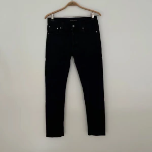 Svarta jeans från Nudie Jeans, W30 L32 - Svarta jeans från Nudie Jeans med klassisk femficksdesign och raka ben. Jeansen har en stilren look med svarta sömmar och diskret logga på bakfickan. Tillverkade i ett mjukt och slitstarkt bomullstyg som sitter snyggt på kroppen.