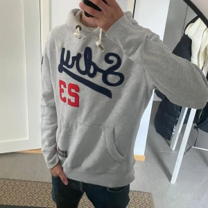 Grå Superdry hoodie med brodyr - Säljer en grå hoodie från Superdry med blå broderad logga och röd siffra 23 på bröstet. Tröjan har huva med snörning, känguruficka och ribbade muddar. Mjuk insida i fleece och snygga detaljer på ärmen och nederkant. Storlek M men skulle kunna passa L