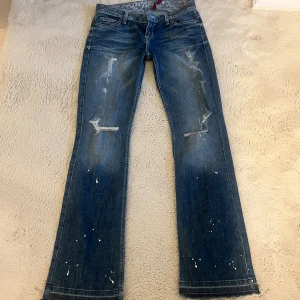 Lågmidjade coola bootcut jeans  - Säljer ett par blå lågmidjade bootcut jeans från GUESS med slitna detaljer samt vita färgstänk som är en del av designen. Fransade benslut.  Innerbenslängd: 83cm Midja: 37cm storlek 27, skulle säga att de passar en XS/ev en liten S 💙
