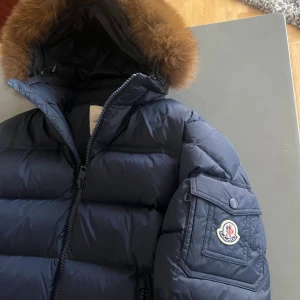 Mörkblå Moncler dunjacka med pälshuva - Snygg mörkblå dunjacka från Moncler med fluffig pälskant på huvan och klassisk logga på ärmen. Jackan har två stora dragkedjefickor framtill, en extra ficka på ärmen och quiltad design. Perfekt för kalla dagar när du vill hålla stilen.