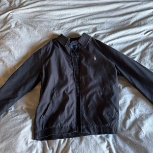 Svart vindjacka från Polo Ralph Lauren - Svart vindjacka (Bi-swing) från Polo Ralph Lauren med klassisk krage, dragkedja och två snedställda fickor. Jackan har broderad logga på bröstet och rutigt foder på insidan. Perfekt för dig som gillar stilren och tidlös design.