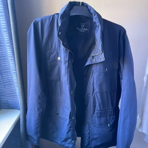 Blå vindjacka från Bondelid, stl M - Snygg marinblå vindjacka från Bondelid med dragkedja och tryckknappar. Jackan har flera praktiska fickor framtill och en hög krage. Materialet är lätt och känns sportigt, perfekt för blåsiga dagar. Klassisk passform och stilren design. Den har en egen utvecklad luva som går att ta fram genom en dragkedja vid nacken. Ny pris kostar 1,800kr men säljer den för 1,300kr för har nästan aldrig använt den.