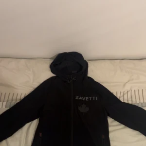 Svart vindjacka från Zavetti - Svart vindjacka från Zavetti med huva och dragkedja framtill. Jackan har tryckt logga och lönnlöv på bröstet samt snörning i huvan. Perfekt för dig som gillar stilren och sportig look.