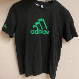 Svart Adidas t-shirt med grön logga - Svart t-shirt från Adidas med grön logga på bröstet och ärmen. Klassisk rund hals och korta ärmar. Tillverkad i mjuk bomull som är skön mot huden. Perfekt för dig som gillar sportig och enkel stil.