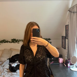 Svart spetsblus från Vila - Svart kortärmad skjorta från Vila i genombruten spets med blommigt mönster. Klassisk krage, knäppning framtill och loose fit. Ärmarna är utsvängda.