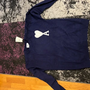 Marinblå stickad tröja från Ami - Säljer en marinblå stickad tröja från Ami Alexandre Mattiussi med deras ikoniska vita hjärta och A-logga på bröstet. Tröjan har rund halsringning, ribbade muddar och är långärmad. Perfekt för dig som vill ha en stilren och trendig look.