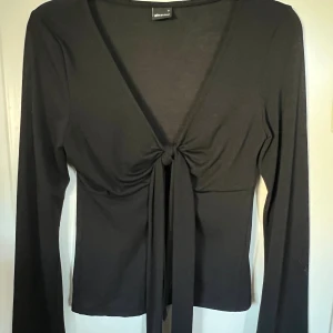 Svart långärmad topp med knyt Gina Tricot - Svart långärmad topp från Gina Tricot med djup v-ringning och snygg knytning framtill. Toppens material är mjukt och stretchigt, vilket gör den bekväm och smickrande. Perfekt att styla med jeans eller kjol för en trendig look.