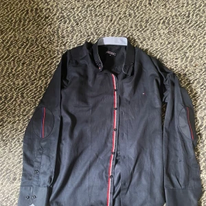 Svart skjorta Tommy Hilfiger M - Svart långärmad skjorta från Tommy Hilfiger i 100% bomull. Snygga detaljer med röd och blå rand längs knappraden och på armbågarna. Klassisk krage och diskret logga på bröstet. Perfekt för dig som gillar stilrena plagg med en twist.