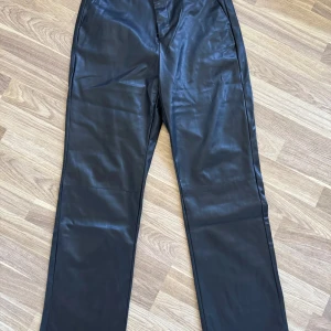 Svarta skinnbyxor från Vero Moda - Snygga svarta byxor i skinnimitation från Vero Moda. De har raka ben, bälteshällor och klassisk passform. Låg/normal midja. Perfekta för dig som vill ha en edgy vibe till din outfit. Materialet är mjukt och ger en cool look.