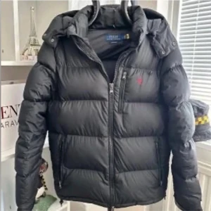 Svart dunjacka från Polo Ralph Lauren - Svart dunjacka från Polo Ralph Lauren med huva och dragkedja. Jackan har en klassisk pufferstil, diskret logga i rött på bröstet och flera fickor med dragkedja. Perfekt för kalla dagar och riktigt snygg till streetwear eller casual outfits.
