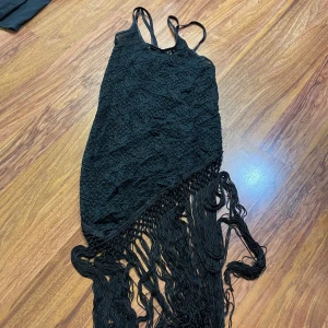 Svart spetsklänning med fransar - Supersnygg svart klänning med smala axelband, spetsdetaljer och långa fransar nedtill. Klänningen har en asymmetrisk nederkant och är täckt av blommig spets, vilket ger en unik och trendig look. Perfekt för dig som vill sticka ut med en cool och elegant stil.