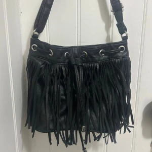 Svart axelväska med fransar - Cool svart axelväska i skinn med långa fransar framtill och snörning upptill. Väskan har en justerbar axelrem och metallöglor som detaljer. Vet inte märke tyvärr då den är köpt för länge sen💓