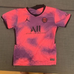 PSG x Jordan kortärmad fotbollströja - Snygg PSG x Jordan fotbollströja med korta ärmar i lila och rosa toner. Tröjan har ett unikt mönster, svart Jordan-logga och PSG-emblem på bröstet. Materialet är lätt och andas, perfekt för träning eller match. Rund hals och svarta detaljer vid sidor och krage.
