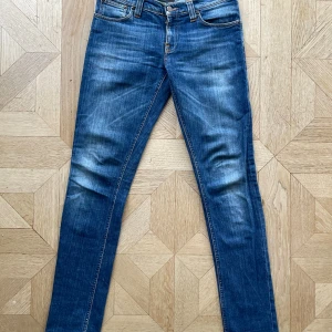Nudie Jeans - Säljer ett par blå skinny jeans från Nudie Jeans med klassisk femficksdesign och snygga slitningar. Jeansen har orangea sömmar, brunt läderpatch med logga och broderade detaljer på bakfickorna. Perfekt passform för dig som gillar smal siluett.