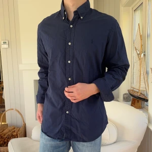 Mörkblå skjorta från Ralph Lauren - Klassisk mörkblå skjorta från Ralph Lauren med button-down krage och vita knappar. Skjortan är i bomull och har lång ärm, perfekt för en clean och stilren look. Diskret broderad logga på bröstet och snygg passform.