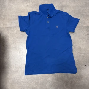 Blå pikétröja från GANT - Snygg blå pikétröja från GANT med klassisk krage och knäppning framtill. Tröjan har korta ärmar och GANT-logga broderad på bröstet. Perfekt för en stilren och sportig look. Tillverkad i mjuk bomull för skön komfort.