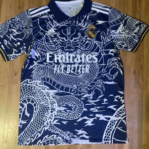 Real Madrid x Adidas blå draktröja - Snygg Real Madrid fotbollströja från Adidas i mörkblått med vit drakdesign över hela tröjan. Klassisk krage, korta ärmar och gula detaljer vid ärmslut. Tillverkad i lätt och ventilerande material, perfekt för match eller träning.