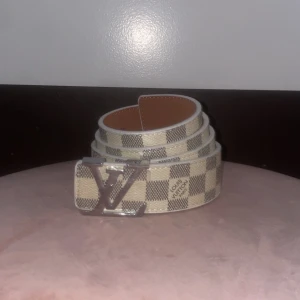 🌟 - Snyggt bälte med klassiskt rutigt mönster i beige och grått. Bältet har en stor silvrig logga som spänne och är gjort i skinn. Perfekt accessoar för att lyfta din outfit.