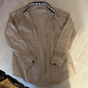 Burberry skjorta - Klassisk beige skjorta från Burberry med ikoniska rutiga detaljer på insidan av kragen och ärmsluten. Skjortan har knappar framtill, lång ärm och diskret broderad logga på bröstet. Tillverkad i mjuk bomull för en clean och stilren look.