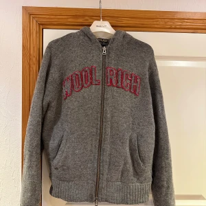 Grå hoodie från Woolrich med dragkedja - Säljer en grå hoodie från Woolrich med röd broderad text på bröstet. Tröjan har huva, hel dragkedja framtill och två fickor. Materialet är mjuk ull och modellen är normal i passningen. Perfekt för dig som gillar chill och stilren look. Ett litet märke på undersidan av armen som ej syns när den används, löses lätt med nål och tråd! 