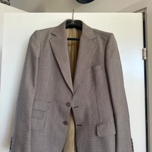 Brunrutig kostym från H&M - Snygg tvådelad kostym från H&M i diskret brunrutigt mönster. Kavajen har klassisk passform med två knappar, spetsiga slag och flera fickor. Byxorna har bälteshällor och bakficka med knapp. Perfekt för dig som gillar stilrena och tidlösa plagg.