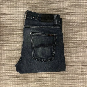Mörkblå jeans från Nudie Jeans - Säljer ett par riktigt feta Nudie Jeans. Osäker på modellen då texten på insidan blekts bort. Skulle uppskatta att det är en av deras smalare modeller såsom ”Grim Tim” eller ”Thin Finn”. Jeansen har ett litet hål på framsidan men är i övrigt i bra skick.                                                      W: 31 L: ~ 32 (Originellt 34, men är uppsydda) ||Topp till botten: ~ 101cm (Uppsydda) || Midjemått: ~ 42cm ||  Lårmått: ~ 26cm  ||  Ankelvidd: ~ 16cm || Hör av er vid frågor eller funderingar!