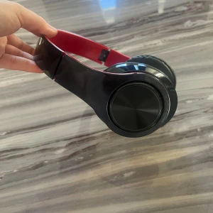 Trådlösa over-ear hörlurar svart/röd - Stilrena trådlösa over-ear hörlurar i svart och rött. Mjuka öronkuddar för bekväm användning. Synliga knappar för volym och kontroll. Inga tydliga märken eller logotyper. Ser ut att vara i gott skick utan synligt slitage.