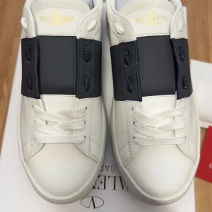 Valentino Garavani Open sneakers vit/blå - Snygga Valentino Garavani Open sneakers i vitt skinn med bred mörkblå rem över snörningen. Klassisk rund tå, platt sula med pyramidmönster och vita skosnören. Guldlogga på plösen och coola detaljer på hälen. Perfekt för dig som gillar exklusiv streetstyle.