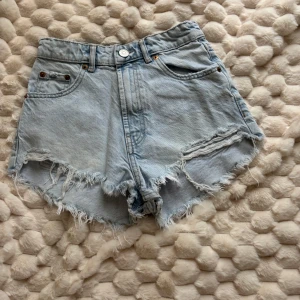 Ljusblå denimshorts från Zara - Säljer ett par ljusblå denimshorts från Zara med hög midja och råa, fransiga kanter. Shortsen har klassiska fem fickor, knapp och dragkedja framtill. Perfekt för sommarens chill dagar och har en loose fit som ger en avslappnad vibe.