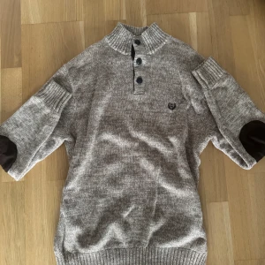 Beige stickad tröja från Chaps - Stickad beige tröja från Chaps med ribbad hög krage och tre knappar framtill. Tröjan har förstärkta armbågar i mörkare tyg och broderad logga på bröstet. Perfekt för kyliga dagar och snygg till jeans.